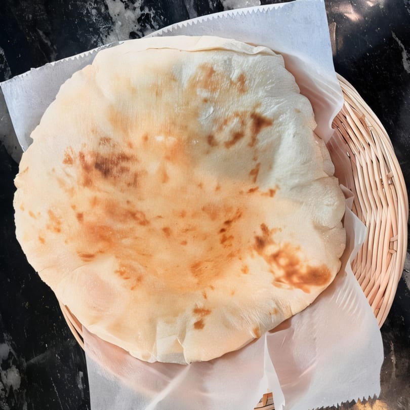 Freshly baked pita bread, lightly browned, served in a woven basket lined with white paper.