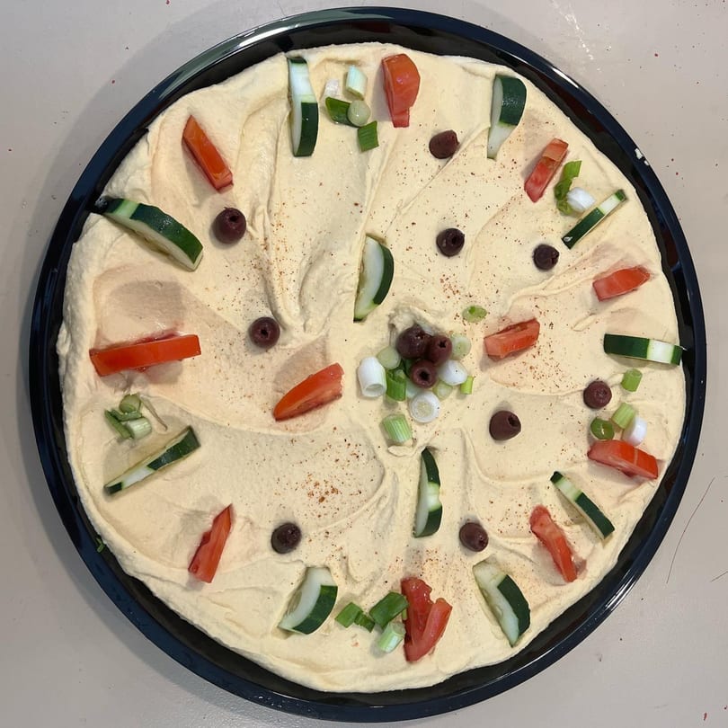 A large platter of Dalia's creamy hummus, garnished with tomatoes, cucumbers, olives, and green onions.